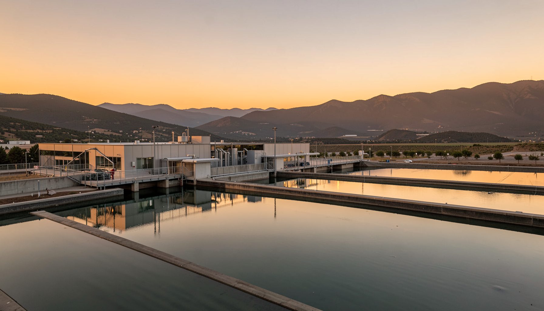 A wide landscape view of a modern water treatment facility at golden hour, with calm reflective water basins stretching across the frame under a warm amber sky