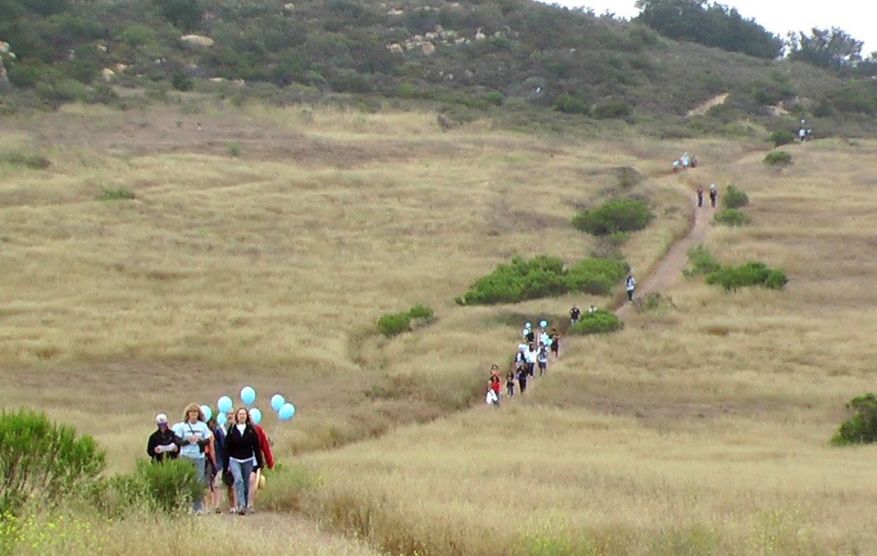 A line of people walking along a winding dirt trail through dry grassy hills, some holding light blue balloons, overcast