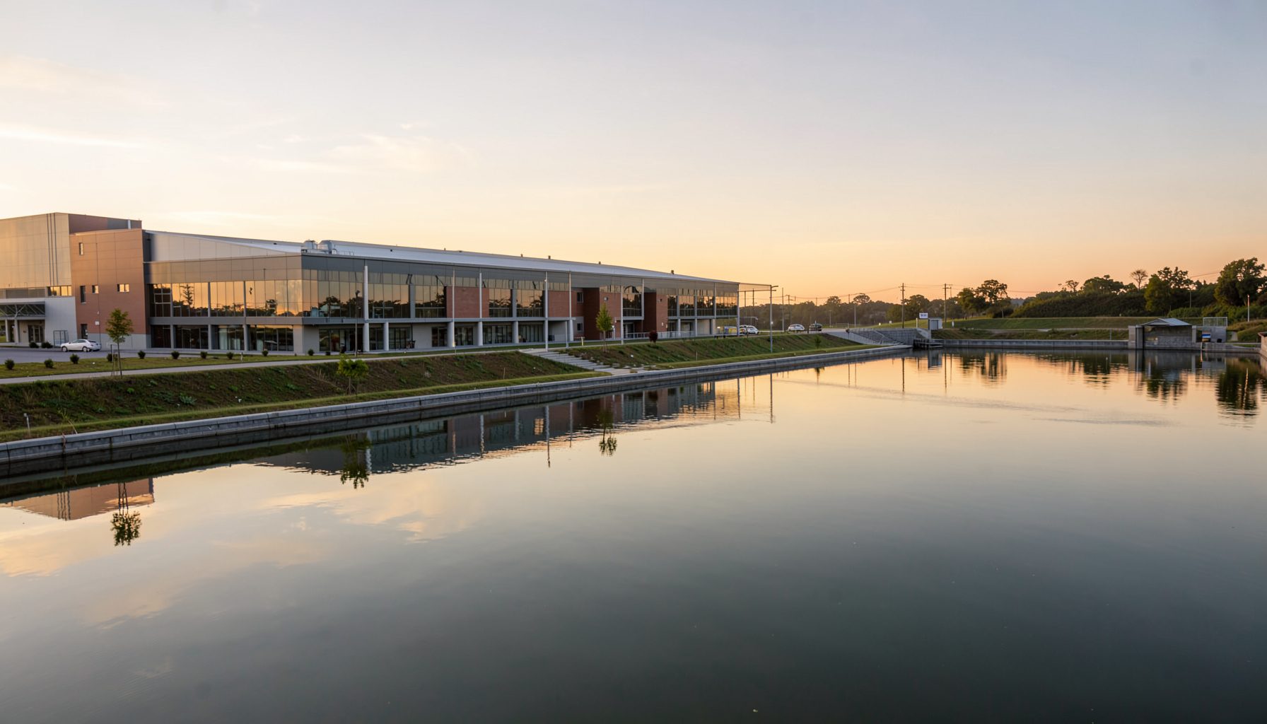 A wide landscape view of a modern water treatment facility at golden hour, with calm reflective water basins stretching across the scene